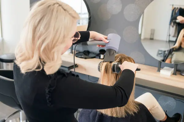 Kadın kuaför stilisti güzellik salonunda çalışıyor ve müşterilere saç stili yapıyor. Sarı saç boyası. Saç kurutuculu kuru saç. Aynaya bakıyor. Sandalyede oturuyor..