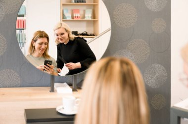 Genç mutlu kadın telefonuna bakıyor, stilistine danışıyor, yeni saç stili ve rengini seçiyor. Kuaför ve müşteri arka plandaki güzellik salonunda günlük kıyafetler giyiyorlar. güzellik ve bakım konsepti