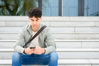 Kendine güveni tam, sıradan kıyafetler giyen çekici bir gencin portresi dışarıda oturuyor, cep telefonu kullanıyor. Genç işadamı akıllı telefon kullanıyor. iş, iletişim konsepti