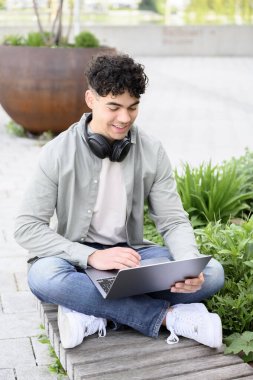 Mutlu öğrenci manzarası, günlük kıyafetlerle serbest çalışan, parkta bankta oturan, dizüstü bilgisayar kullanan, güneşli bir bahar gününde çalışan,.