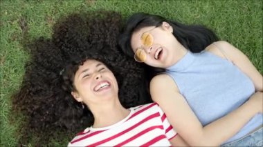 İki genç bayan arkadaş çimlerin üzerinde yatıyor, biri çizgili gömlek giyiyor ve diğeri güneş gözlüğü takıyor. Gülümsüyorlar, konuşuyorlar, birlikte eğleniyorlar.