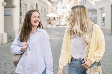 Kafkasyalı güzel, uzun sarı ve esmer saçlı, günlük giysiler içinde Avrupa 'da bir yaya caddesinde yürüyen bir şehirde konuşan ve gülümseyen iki kadın. yaşam tarzı konsepti