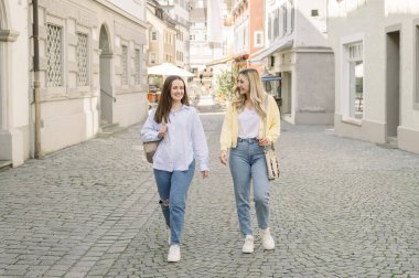 24-29 yaşlarında, kot pantolon, gömlek, ceket giymiş, güneşli bir günde Avrupa 'da şehir merkezini ziyaret eden yaya caddesinde yürüyen iki beyaz kadın arkadaş. seyahat ve yaşam tarzı kavramı