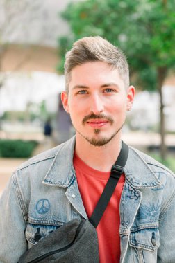 Sakallı, kırmızı tişörtlü, kot ceket giyen, siyah omuz çantalı, kameraya bakan pozitif bir gencin portresi. yaşam tarzı konsepti