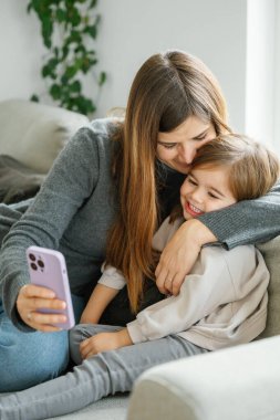 Genç, sevgi dolu bir anne evde küçük kızına sarılıyor ve kameraya gülümsüyor, koşulsuz sevgisini ifade ediyor. Anne-kız ilişkisi, mutlu annelik, ergenlik çağında ebeveynlik..