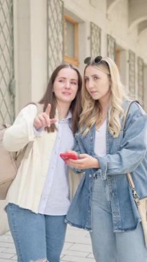 Sırt çantalı ve bavullu iki kadın turistin şehirde yol tarifi almak için cep telefonu kullanırken çekilmiş dikey fotoğrafı. seyahat kavramı