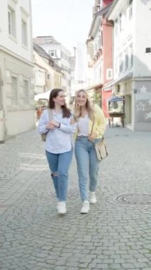 24-29 yaşlarında, kot pantolon, gömlek, ceket giymiş, güneşli bir günde Avrupa 'da şehir merkezini ziyaret eden yaya caddesinde yürüyen iki beyaz kadın arkadaş. seyahat ve yaşam tarzı kavramı