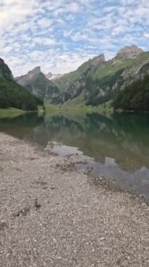 Güneşli bir günde arka planda dağ kulübeleriyle yeşil bir vadide dağlık bir gölde yürüyen turistlerin panoramik görüntüsü. Seealpsee, Wasserauen, Appenzell, İsviçre.