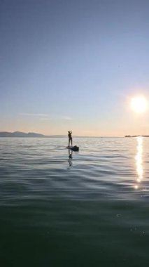 40 'larında kısa boylu ve bikinili bir kadın Almanya' da Bodensee 'de bir gölde ayakta duran kürek tahtasının üzerinde duruyor, mavi gökyüzü pürüzsüz suya yansıyor, arka planda güneş batıyor, günbatımı.