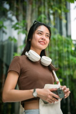 Elinde cep telefonu tutan genç bir Asyalı kadın, günlük kıyafetler giyiyor, çanta, kulaklık takıyor, Bangkok, Tayland 'da bir şehir ortamında rahat görünüyor.
