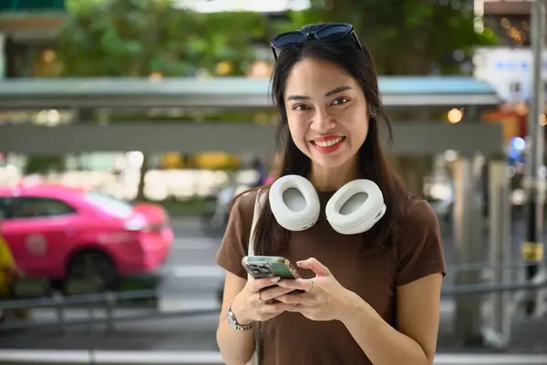 Kulaklıkları ve çantasıyla mutlu bir Asyalı kadın hafta sonu kaçamağı sırasında Bangkok Tayland 'da şehir gezisinin tadını çıkarırken akıllı telefonunu kullanırken gülümsüyor. seyahat kavramı