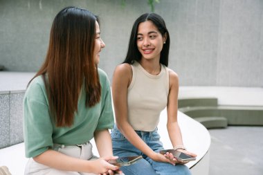 Sıradan kıyafetler içinde iki genç Asyalı kadın, Bangkok 'ta modern bir açık hava ortamında oturup konuşuyorlar, gülümsüyorlar ve ellerinde akıllı telefonlarla birbirleriyle eğleniyorlar. yaşam tarzı konsepti