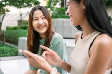 İki genç Asyalı kadın nedensel kıyafetler içinde, bir kahvenin dışında oturuyor, dışarıda keyifli bir anı paylaşıyor, akıllı bir telefonla konuşurken birlikte gülüyorlar. yaşam tarzı konsepti