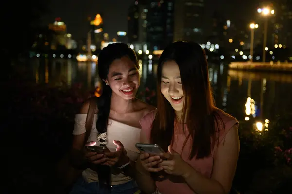 İki genç Taylandlı kadın, telefon ekranlarıyla aydınlatılmış, nehir kenarında sakin bir akşamın tadını çıkarıyorlar. Parlak Bangkok silueti arka fon olarak, gülümsüyor, konuşuyor. arkadaşlık ve yaşam tarzı kavramı