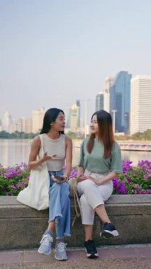 İki Asyalı kız arkadaş, gündelik kıyafetlerle parkta oturup sohbet ediyorlar, birlikte eğleniyorlar hafta sonları şehir gezisi sırasında Bangkok 'ta vakit geçiriyorlar. seyahat ve yaşam tarzı kavramı