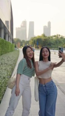 Taylandlı iki genç kadın Bangkok 'ta keyifli bir anın tadını çıkarıyorlar. Canlı bir şehir parkında, arka plandaki Bangkok siluetinde selfie çekiyorlar. yaşam tarzı konsepti