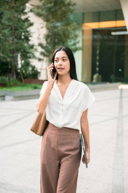 Asyalı genç bir kadın profesyonel, iş kıyafeti giymiş, uzun saçlı, şehir ortamında yürüyen, elinde tablet olan, Bangkok Tayland 'da bir binanın önünde akıllı telefondan konuşan