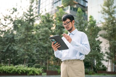 Açık havada tablet kullanan profesyonel bir adam, çevresi yeşillik ve modern mimariyle çevrili, görevine odaklanıyor.