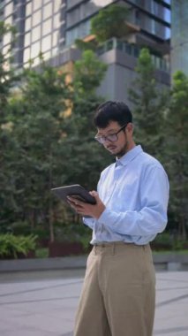 Gözlük takan, dışarıda tablet kullanan, yeşillik ve modern mimari ile çevrili, Bangkok 'taki işine odaklanan profesyonel iş adamı.
