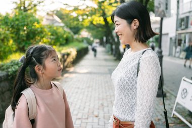 Mutlu Asyalı öğrenci ve annesi günlük giysiler içinde, Japonya 'da okuldan eve yürüyerek dönüyorlar, sohbet ediyorlar ve gülümsüyorlar, aile, eğitim ve çocukluk sevincini şehir ortamında yansıtıyorlar. aile kavramı