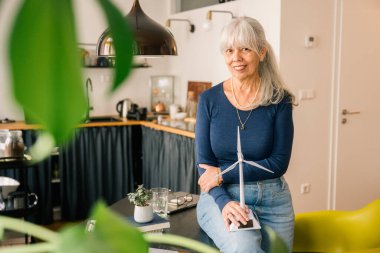 Mutfak tezgahında oturan gülümseyen yaşlı kadın, elinde küçük bir rüzgar türbini modeli tutuyor ve konut ortamlarında yenilenebilir enerji ve sürdürülebilir yaşam tarzının kullanımını teşvik ediyor. çevre kavramı