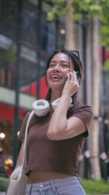 Boynunda kulaklıkları ve omzunda tekrar kullanılabilir bir alışveriş çantası olan neşeli genç Tayland 'da yürürken akıllı telefonuyla mutlu bir şekilde konuşan Taylandlı kadın. yaşam tarzı konsepti