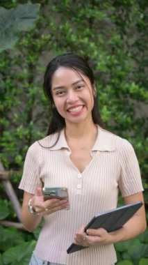 Kendine güvenen Taylandlı iş kadını dışarıda duruyor, elinde akıllı telefon ve tabletle, yemyeşil yeşil manzaranın ortasında mesleki başarı yayıyor.