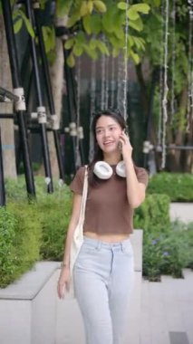 Sıradan kıyafetli şık genç Taylandlı kadın Bangkok 'taki modern bir şehir parkında yürüyor, çantasıyla ve boynunda kulaklıkla telefon konuşmalarından hoşlanıyor. yaşam tarzı konsepti