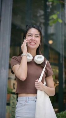 Boynunda kulaklıkları ve omzunda tekrar kullanılabilir bir alışveriş çantası olan neşeli genç Tayland 'da yürürken akıllı telefonuyla mutlu bir şekilde konuşan Taylandlı kadın. yaşam tarzı konsepti