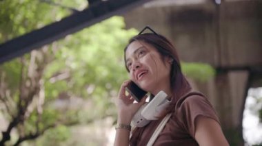 Genç Taylandlı kadın güneş gözlüğü takıyor ve kulaklık takıyor. Cep telefonuyla konuşurken gülümsüyor. Bangkok sokaklarında arka planda yeşillikle dikiliyor.