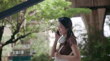 Yeniden kullanılabilir alışveriş çantasıyla neşeli genç Taylandlı kadın Bangkok 'ta üst geçidin altında yürürken telefonuyla hoş bir sohbet ediyor.