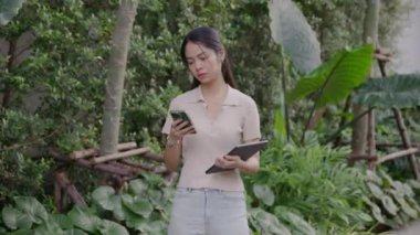 Genç Taylandlı iş kadını yemyeşil bahçede duruyor, telefonla konuşurken tableti dengeliyor, doğal ortamda profesyonel sakinliği somutlaştırıyor.