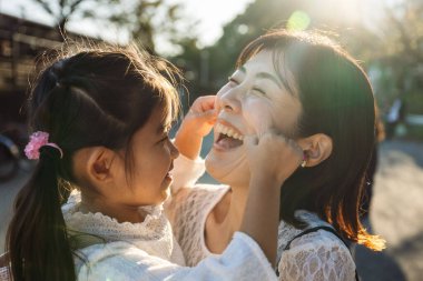 Neşeli Asyalı kızı neşeli bir şekilde neşeli annelerinin yanaklarını açık havada çeker, güneşli bir günde, parkta birbirlerine eşlik ederek eğlenirler. Ebeveynlik kavramı
