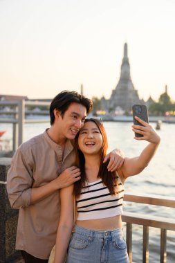 Genç Asyalı turistler Bangkok 'ta tatillerinin tadını çıkarıyorlar. Arka planda ünlü Wat Arun tapınağıyla selfie çekiyorlar. Gün batımında unutulmaz bir anı yakalıyorlar.