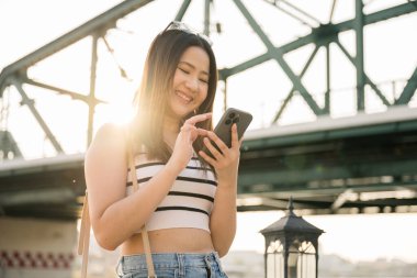 Akıllı telefon kullanarak gülümseyen genç bir kadın, arka planda bir köprüyle dışarıya mesaj gönderiyor. Güneş gün batımında sıcak ve canlı bir atmosfer yaratarak ışıldıyor. İnsanlar, yaşam tarzı konsepti
