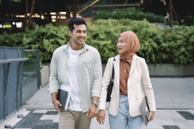 İki genç profesyonel, tesettür giyen bir erkek ve bir kadın, modern bir şehir ortamında dışarıda yürüyorlar, dizüstü bilgisayar taşıyorlar ve sohbet ediyorlar.