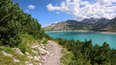 Luenersee gölünün turkuaz suyu yeşil yamaçlar ve kayalık dağlarla çevrili beyaz bulutlu mavi gökyüzünü parıldatarak Avusturya Alpleri 'ndeki yürüyüş yolu boyunca çarpıcı manzaralar sunuyor. Doğa kavramı