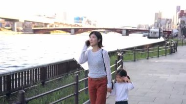 Japon kadın kızıyla dışarıda yürüyor, cep telefonu tutuyor, telefon görüşmesi yapıyor, Tokyo, Japonya 'daki nehirde birlikte vakit geçirirken sohbet ediyor. insanlar kavramı