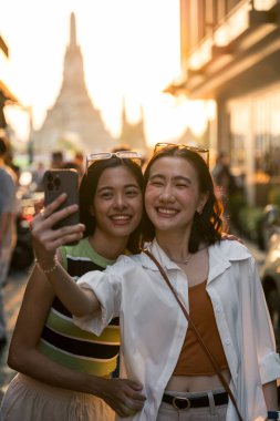 Gülümseyen, selfie çeken iki neşeli bayan turist Bangkok sokaklarında akıllı bir telefonla fotoğraf gönderiyorlar. Tayland. Gün batımında arka planda Wat Arun Tapınağı var. seyahat kavramı