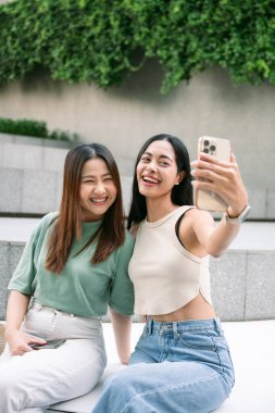 Bangkok, Tayland 'da bir bankta oturan iki neşeli genç kadın, akıllı telefonlarıyla eğlenceli bir selfie çekiyorlar, unutulmaz tatillerinde birlikte kahkaha ve mutluluk paylaşıyorlar.