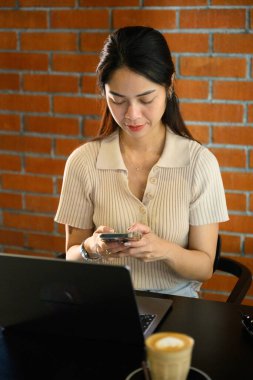 Genç Asyalı serbest yazar, uzun saçlı girişimci kadın, oturuyor, Bangkok 'ta modaya uygun bir kafede çalışırken akıllı telefon kullanıyor, dizüstü bilgisayarı ve kapuçinosu var. iş kavramı