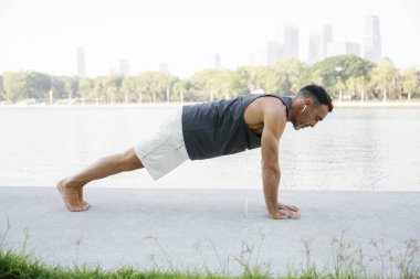 Spor kıyafetleri giyen olgun bir adam, şehir parkında bir gölün kenarında tahta bir poz tutuyor. Açık hava antrenmanı sırasında güç ve dengeyi gösteriyor. spor yaşam tarzı konsepti