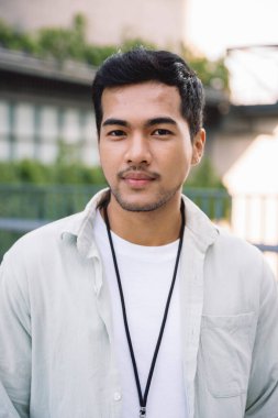 Kısa siyah saçlı, açık yeşil gömlekli, beyaz tişörtlü, dışarıda bulanık bir şehir ortamında duran Asyalı genç bir adamın portresi.