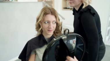 Sarışın bir kadın saçını bir modacıya yaptırıyor. Stilist müşteriye saçlarını aynada gösteriyor, kadın gülümsüyor, sonuçtan memnun olduğunu gösteriyor. güzellik ve bakım konsepti