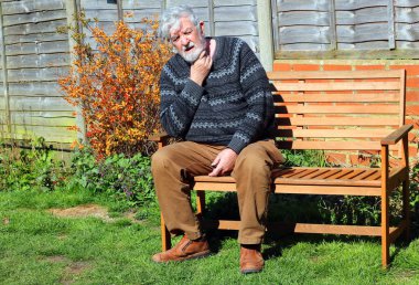 Yaşlı ya da yaşlı bir adam bankta oturup boğazını tutuyor. Boğazı, enfeksiyonu ya da virüsü var. Hasta ve keyifsiz hissediyorum..