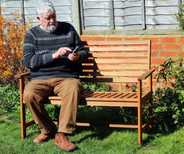 Yaşlı ya da yaşlı bir adam parkta bankta oturuyor ve cep telefonuna veya cep telefonuna mesaj yolluyor..