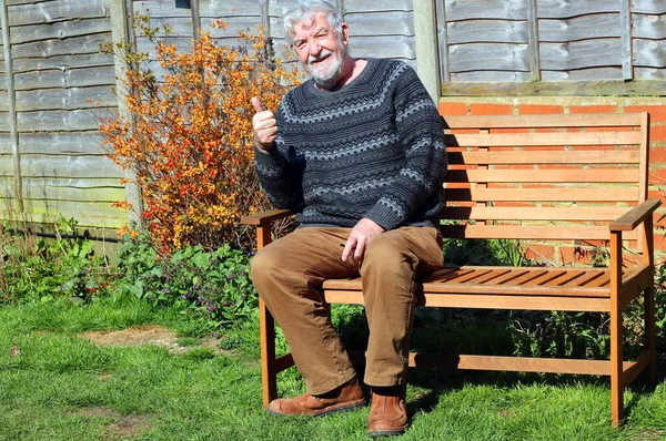Happy elderly or senior man sitting on a bench in his garden and looking towards the camera. Smiling and raising his thumb to say all is fine. Happily retired and healthy.