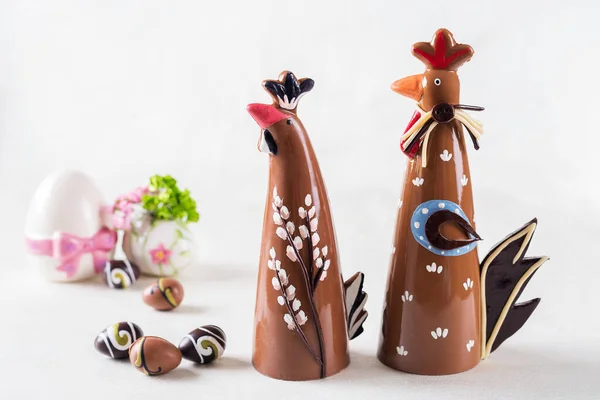 Etrafında çikolata yumurtaları olan beyaz bir tabakta iki çikolatalı horoz.