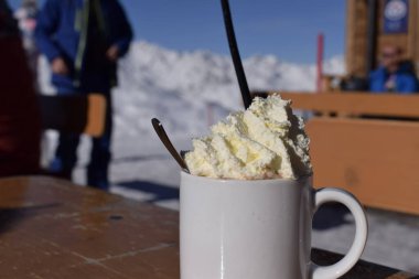 Karlı kayak merkezinde nefis sıcak çikolata. Beyaz kupada, kar kaplı dağ zeminli yumuşak krem şanti. Kayak ve snowboard tatili sırasında dinlenmek için ideal sıcak içecek.