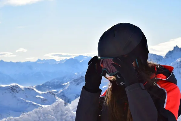 Female skier in winter sports gear with long hair, a black helmet, orange goggles and black face mask. Backdropped by snowcapped Italian mountain peaks of the Pennine alps on a clear, sunny day.